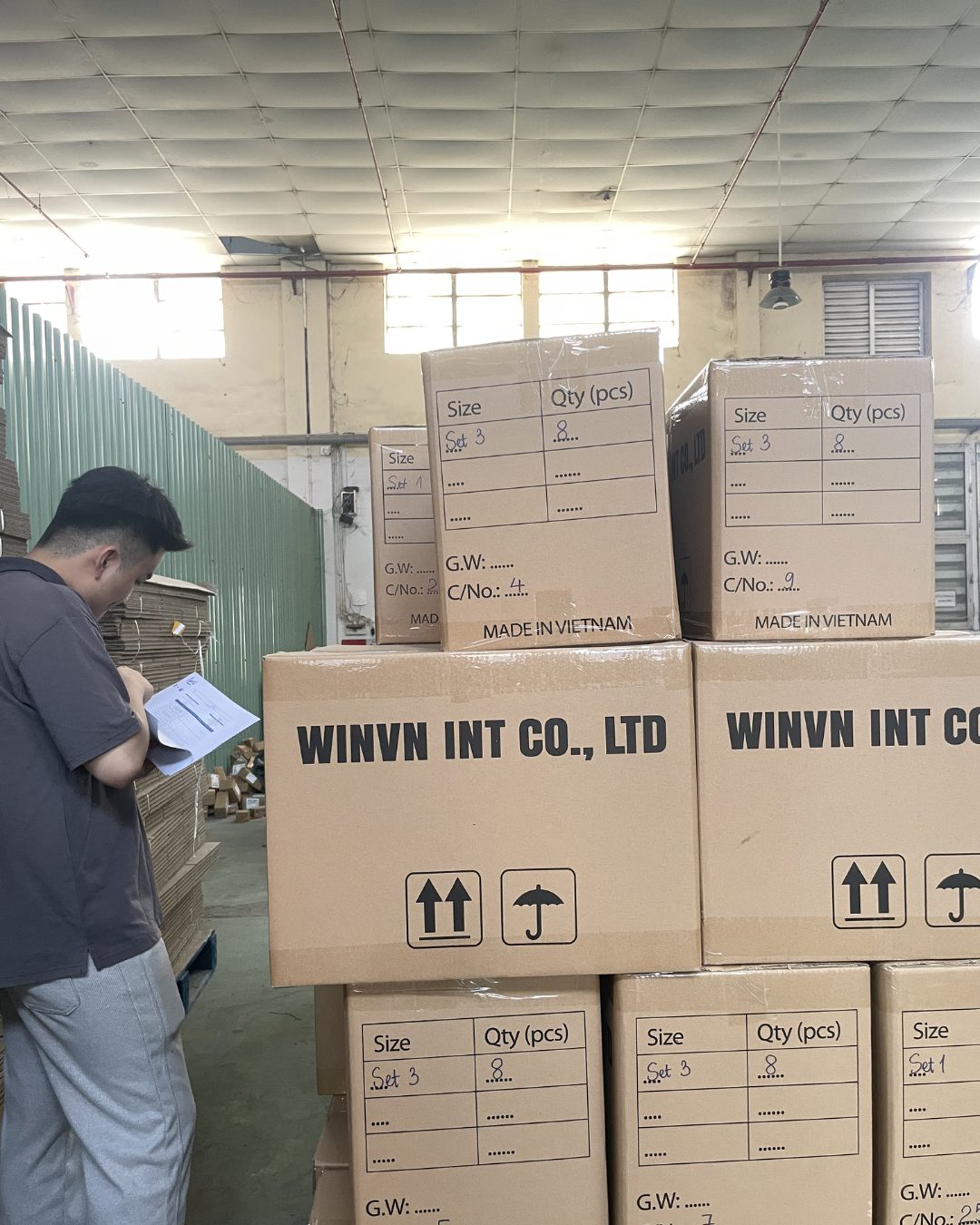 Staff member with a clipboard checking stacked export cartons whose printed panels show size, quantity, gross weight and carton number